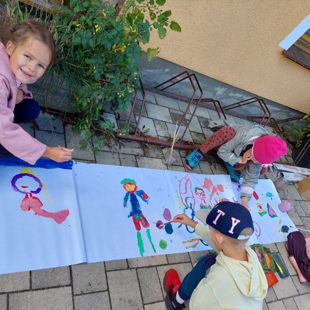 Dzieci malują farbami na arkuszu papieru, który rozłożony jest na ziemi