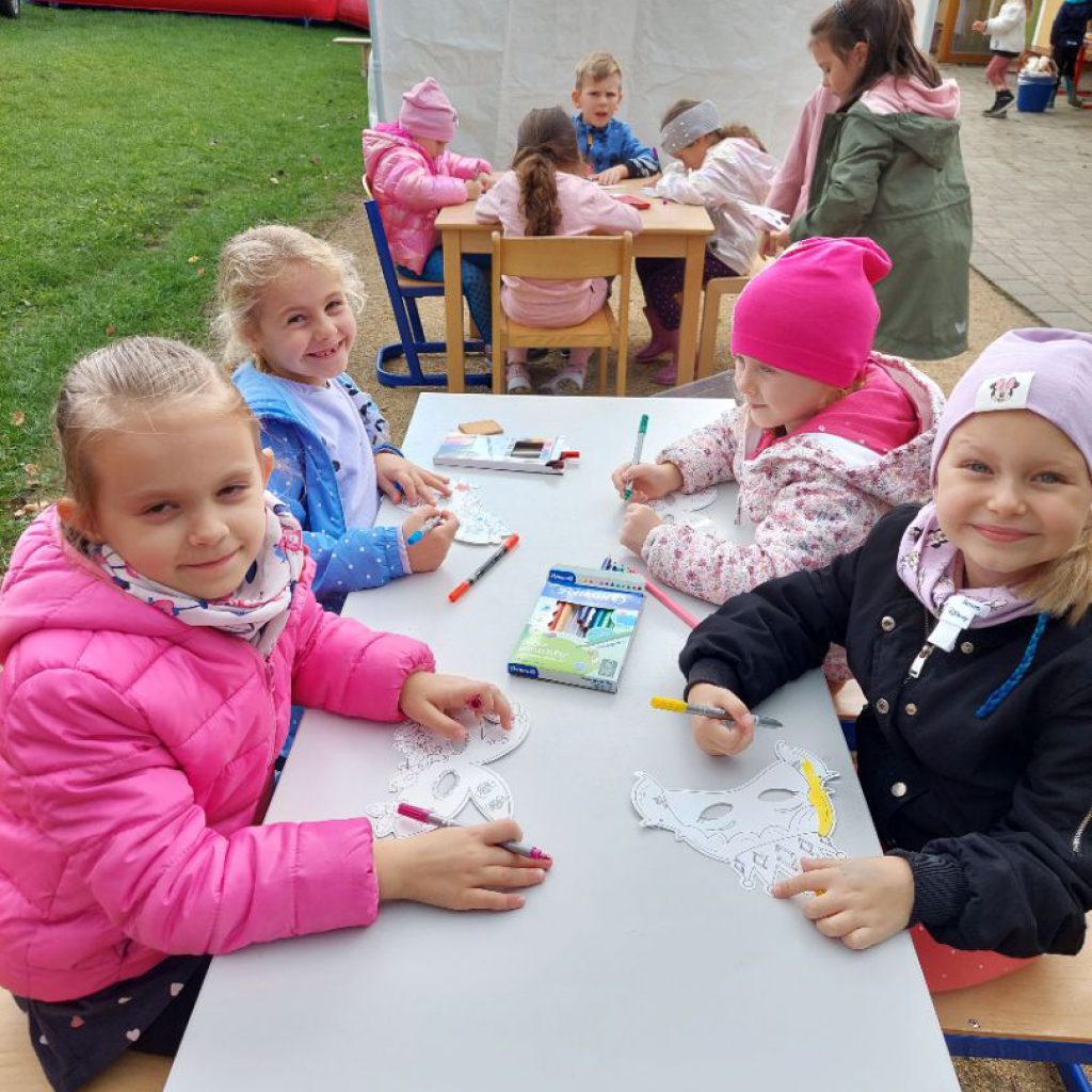 Dzieci siedząc przy stolikach kolorują mazakami maski wykonane z papieru