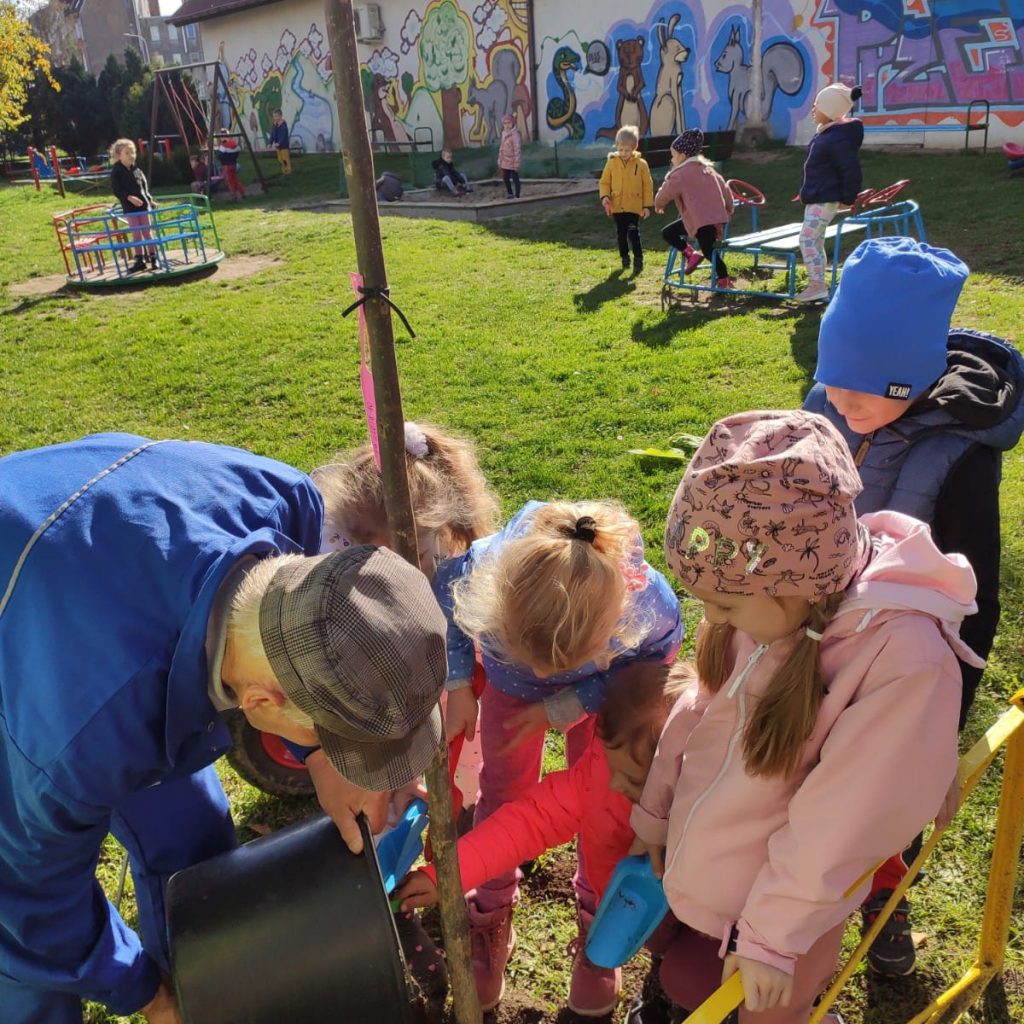 Dzieci z panem konserwatorem sadzą drzewo na placu przedszkolnym