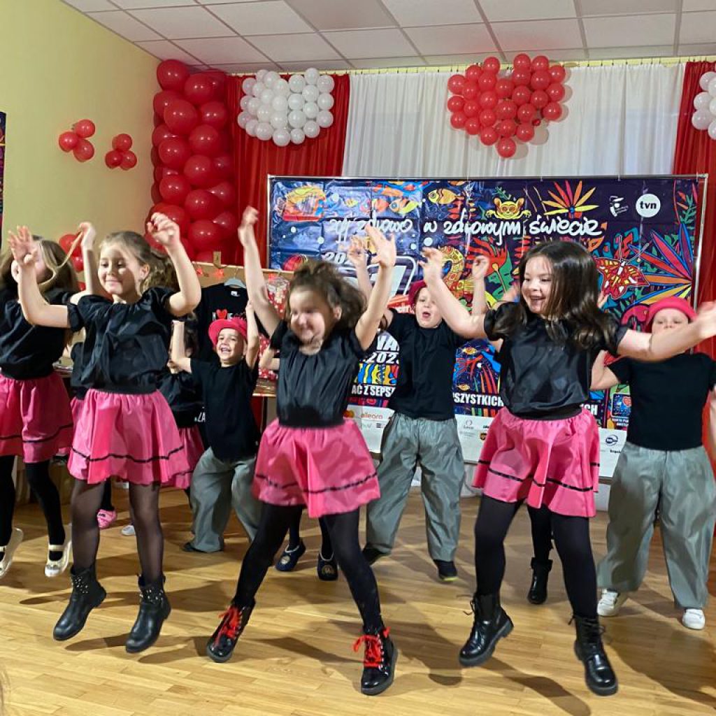 grupa dzieci w różowo czarnych strojach tańczy na środku zaaranżowanej sceny