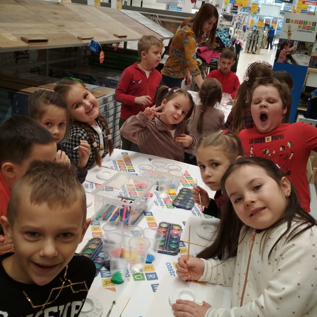 Dzieci pozują przy stolikach do zdjęcia robiąc zabawne miny w trakcie malowania świeczników przy stołach 