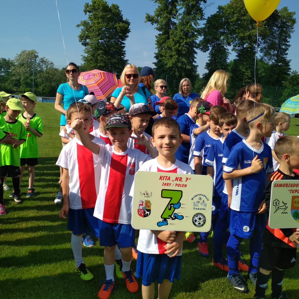 Dzieci ustawione w rzędy, ubrane w stroje sportowe, każdy reprezentant drużyny trzyma w ręku tabliczkę z nazwą przedszkola, z którego przyjechał