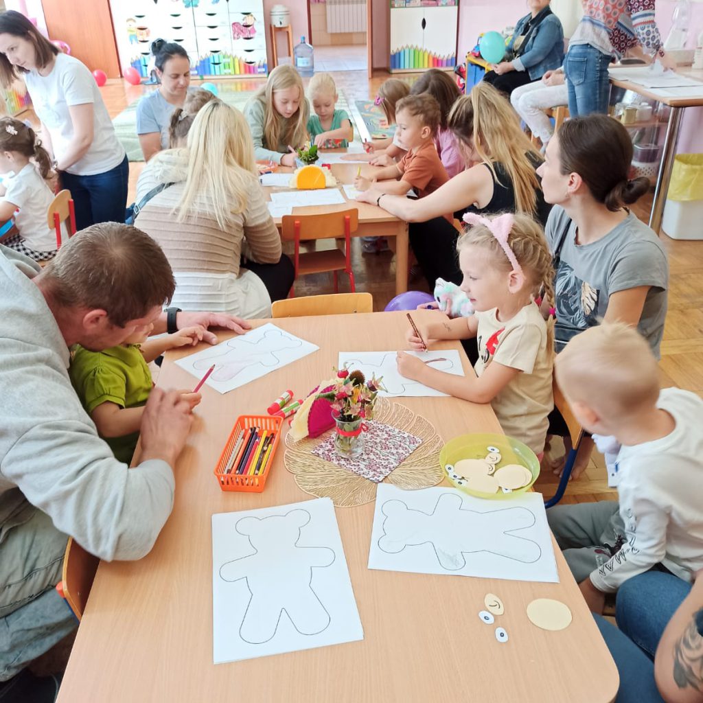 Dzieci wraz z rodzicami siedzą przy stolikach i kolorują szablon misia.