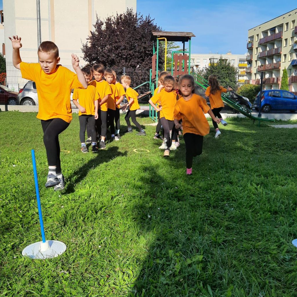 Dzieci ubrane w żółte koszulki bawią się w slalom na trawie.
