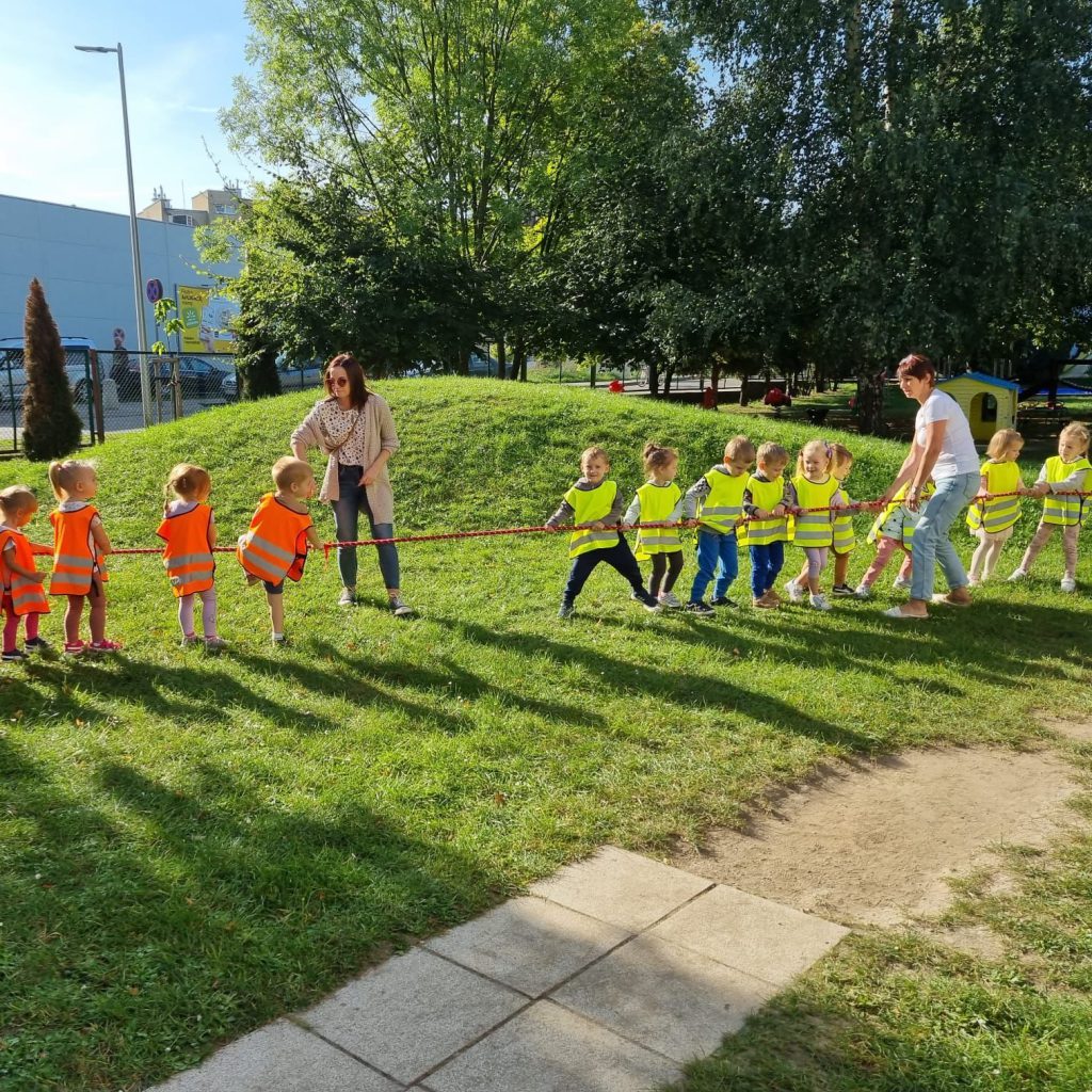 Dzieci z grup maluszków bawią się w przeciąganie liny na trawie.