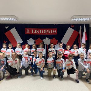 Dzieci ubrane w barwach narodowych, stoją na tle flagi Polski.