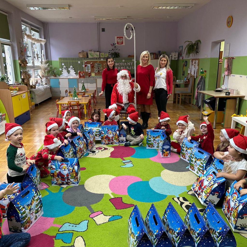 grupa dzieci wraz z opiekunkami i świętym Mikołąjem pozuje w sali siedząc na dywanie