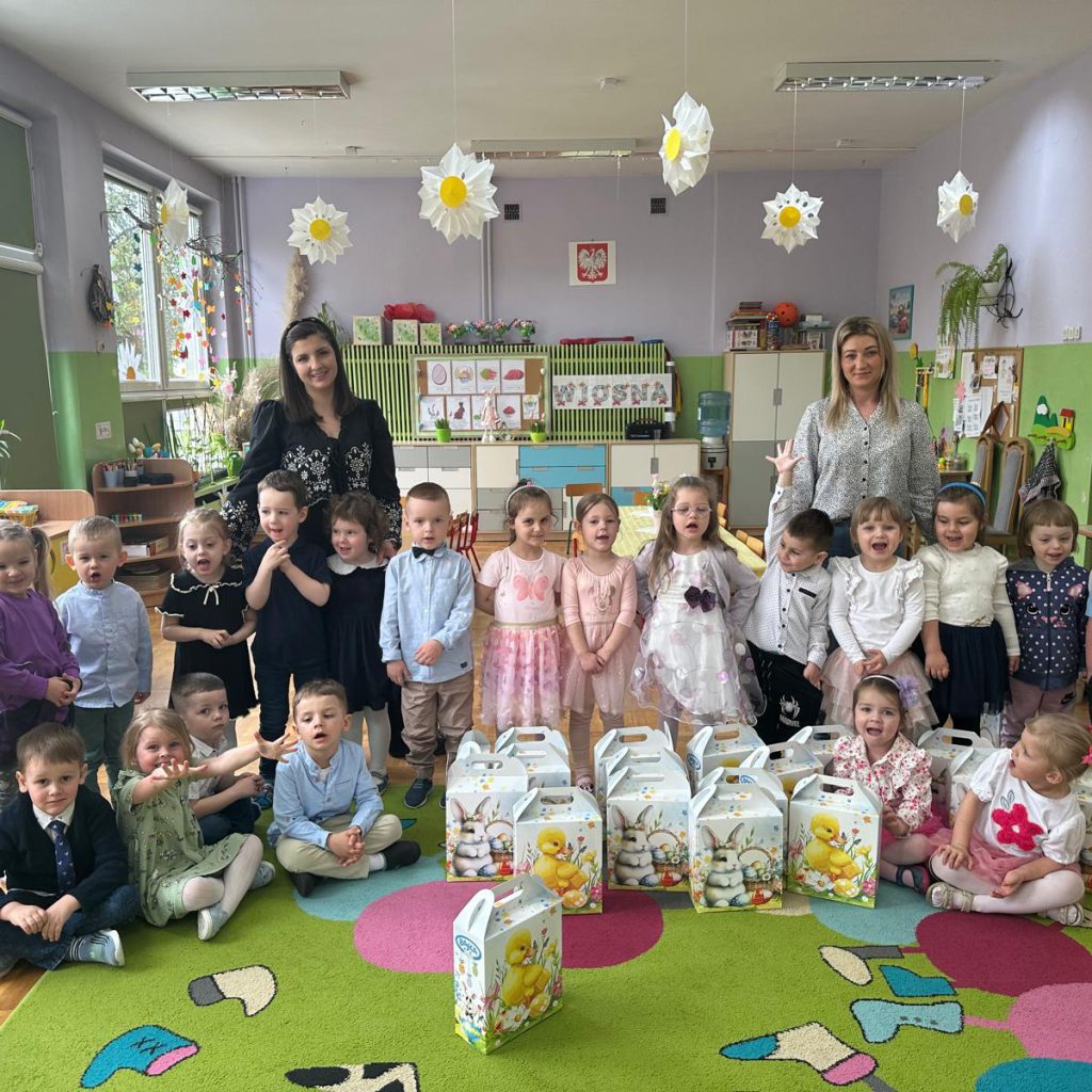 grupa dzieci siedzi na dywanie, a przed nimi znajdują się prezenty od Zająca