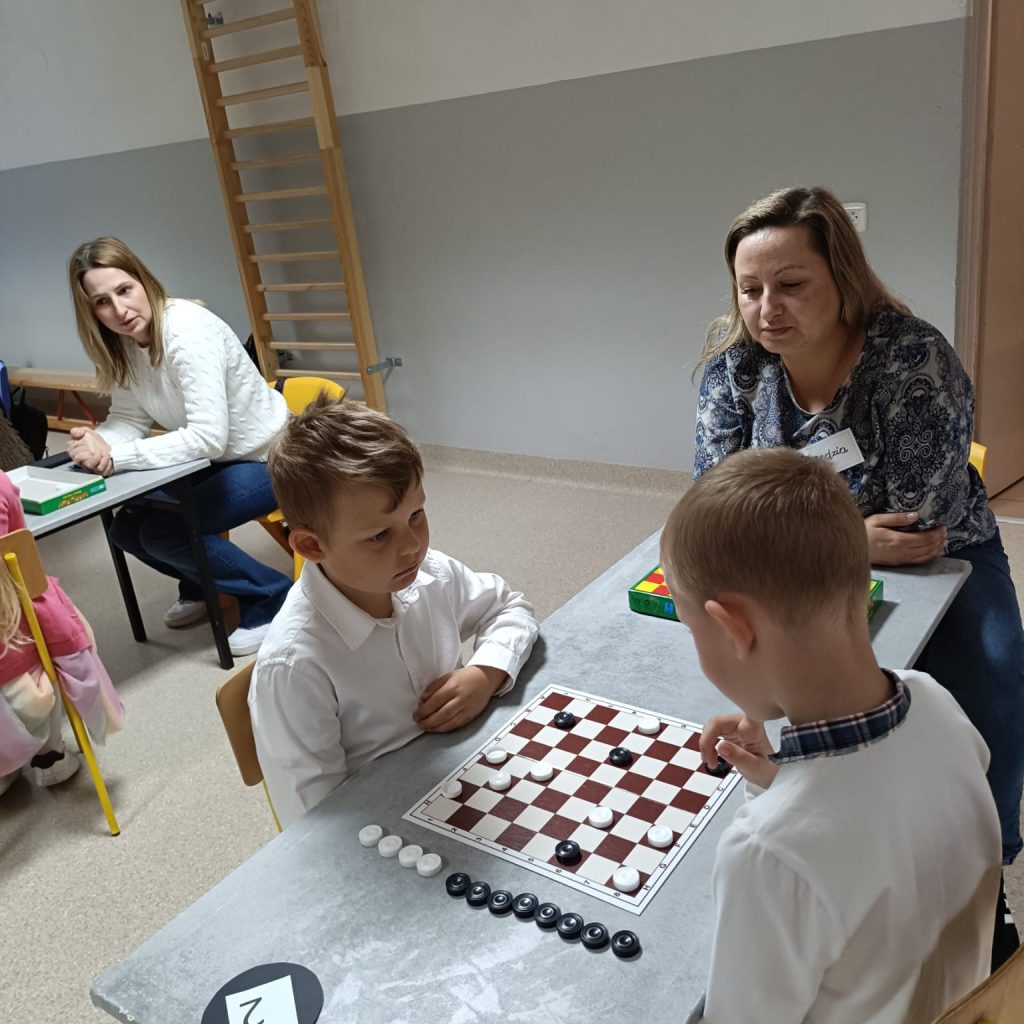 dwoje dzieci i dorosłych gra i obserwuje grę w warcaby