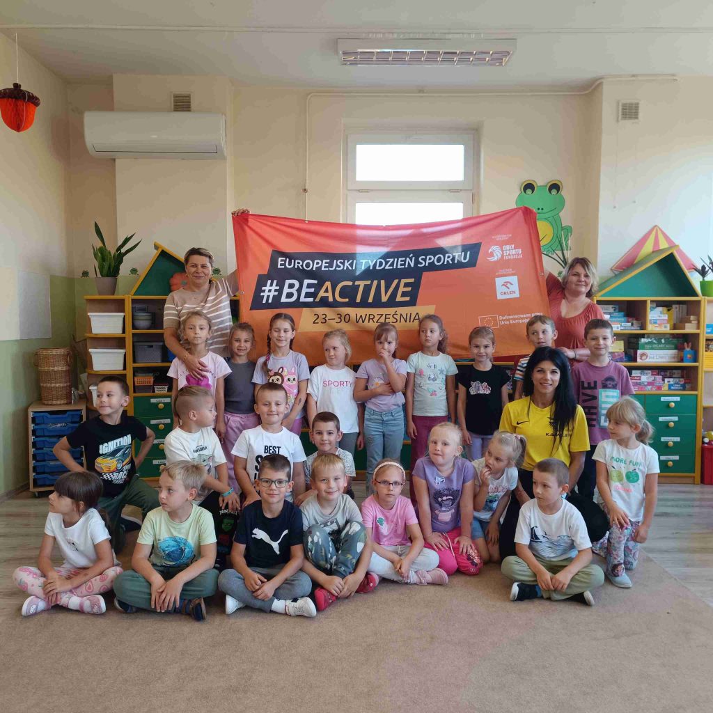 dzieci pozują wraz z opiekunami na tle banera #beactive