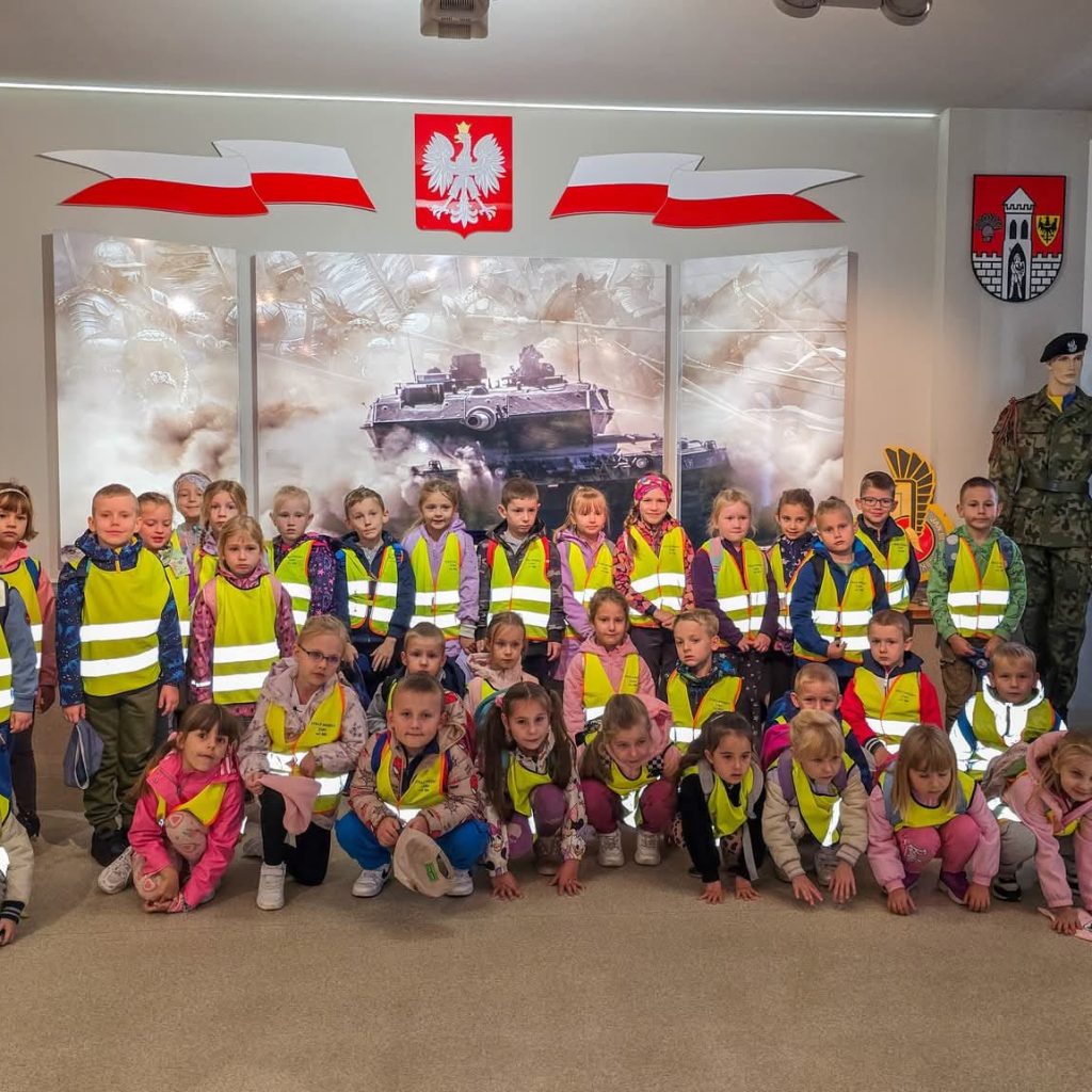dzieci w kamizelkach odblaskowych pozują na tle obrazu przedstawiającego czołg