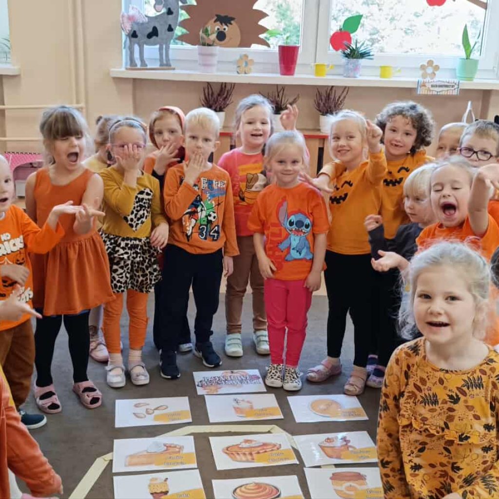 grupa dzieci ubranych na pomarańczowo stoi dookoła prac plastycznych