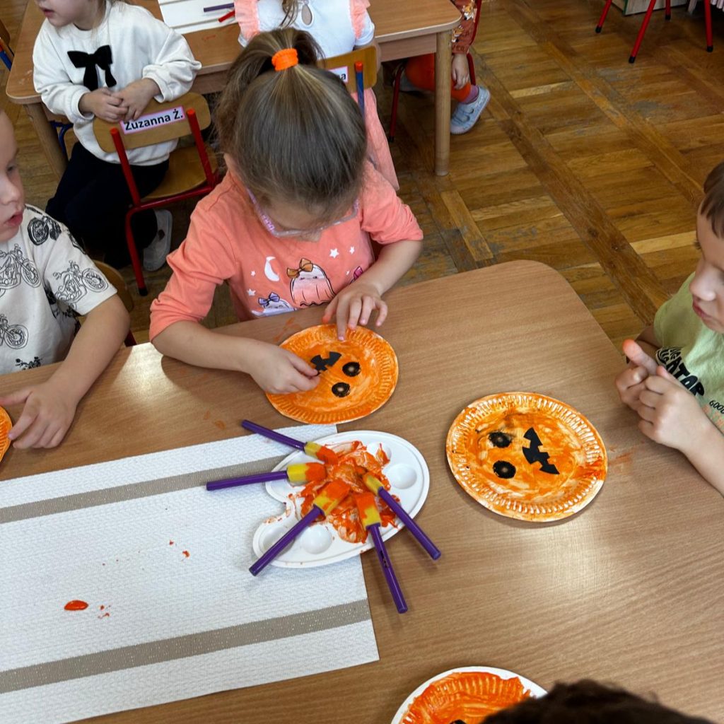 dzieci malują przy stoliku pomarańczową farbą talerze papierowe